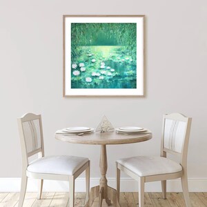 Puede incluir: Una pintura enmarcada de un estanque verde con nen&uacute;fares blancos. El estanque est&aacute; rodeado de &aacute;rboles verdes y el agua refleja la luz del sol. La pintura est&aacute; colgada en una pared blanca en un comedor con dos sillas y una mesa.