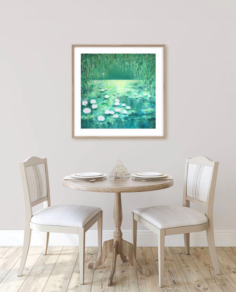 Puede incluir: Una pintura enmarcada de un estanque verde con nen&uacute;fares blancos. El estanque est&aacute; rodeado de &aacute;rboles verdes y el agua refleja la luz del sol. La pintura est&aacute; colgada en una pared blanca en un comedor con dos sillas y una mesa.