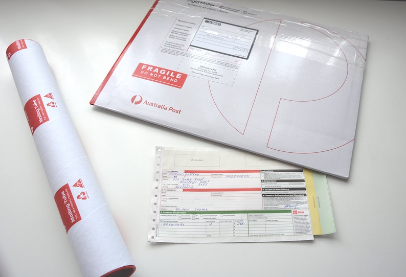 May include: A white mailing tube with red text and a recycling symbol. The tube is rolled up and has a red edge. A white envelope with red text that says "Fragile Do Not Bend" and "Australia Post" is lying flat on a white surface. A white sheet of paper with handwritten text and a red bar code is also on the surface.