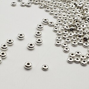 May include: A large quantity of silver-toned metal beads, scattered on a white surface. The beads are round with a hole in the center. Some beads are grouped together in small piles.