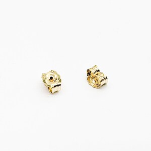 May include: Two gold-colored earring backs on a white background. The earring backs are small, with a butterfly-shaped design. The backs are likely made of metal and are used to secure earrings.