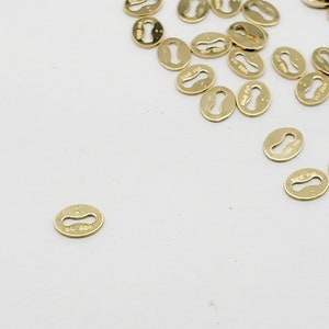 May include: A group of gold-colored metal keyhole-shaped charms. The charms are scattered on a white surface.