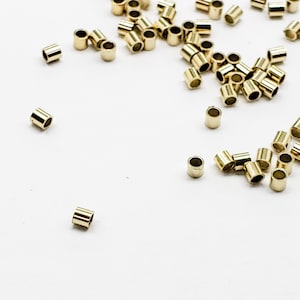 May include: A scattering of gold-colored metal crimp beads on a white surface. The beads are small and cylindrical in shape.