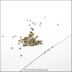 May include: A pile of gold-colored metal crimp beads on a white surface. There are also a few beads strung on a white thread.