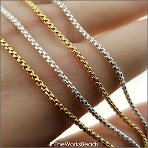 Könnte beinhalten: Drei Kettenstränge, einer in Gold, einer in Silber und einer in Weißgold, werden auf einer Hand gezeigt. Die Ketten sind alle im gleichen Stil, mit kleinen, runden Gliedern.