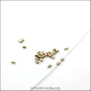 May include: A close-up of a pile of gold-colored crimp beads, some of which are strung on a white thread. The beads are small and cylindrical in shape.