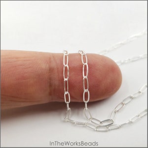 May include: Two silver oval chain necklaces, one is slightly thicker than the other, are shown on a finger. The chains are made of small, oval-shaped links.