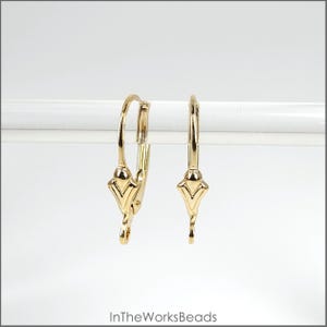 May include: Two gold-colored leverback earrings. Each earring features a decorative, stylized design at the bottom and a curved lever. The earrings are displayed against a white background.