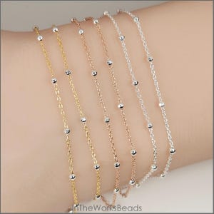 May include: Close-up of several delicate chain bracelets. Each bracelet has small, evenly spaced silver beads. The chains are in gold, rose gold, and silver tones. TheWorksBeads is printed at the bottom.