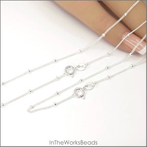 May include: Two silver bead chains with lobster clasps. The chains are layered on top of each other and are shown on a hand.