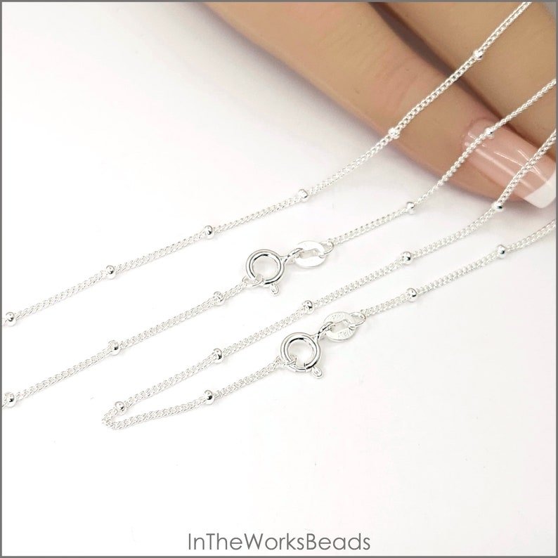 May include: Two silver bead chains with lobster clasps. The chains are layered on top of each other and are shown on a hand.