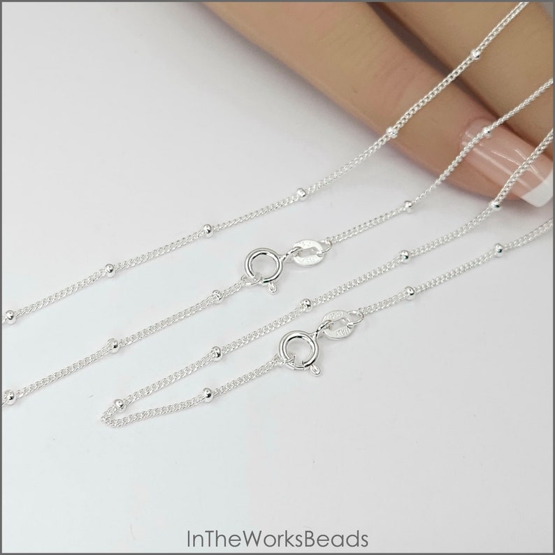 May include: Two silver bead chains with lobster clasps. The chains are laid out on a white surface.