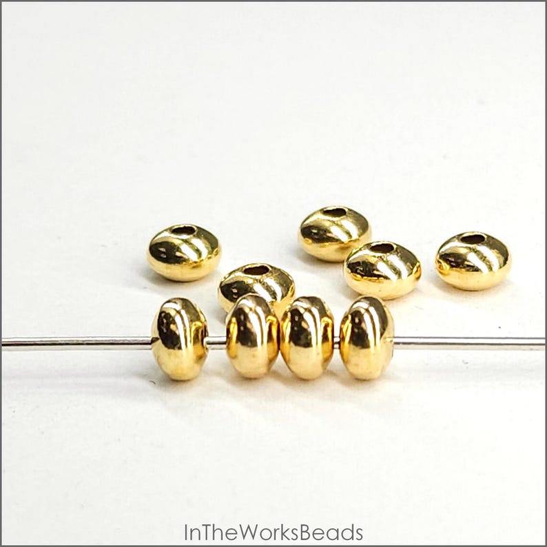 May include: Gold-coloured metal beads with a smooth, rounded surface. The beads are strung on a thin wire and are arranged in a row. There are also several loose beads scattered around the wire.