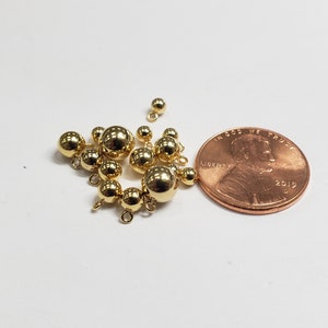 May include: A group of gold-colored metal beads with loops, some of which are connected to each other. The beads are arranged in a loose cluster, and a US penny is included for scale.