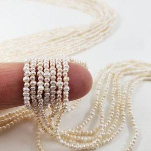 May include: A strand of white freshwater pearls, strung on a thin thread, is shown in a close-up image. The pearls are small and uniform in size, and they are arranged in a neat, even row. The pearls are a classic and elegant choice for jewelry making.