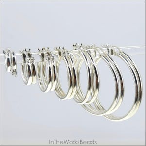 Puede incluir: Un conjunto de pendientes de aro de plata en varios tamaños. Los aros son todos lisos y brillantes, y están dispuestos en una fila del más pequeño al más grande.