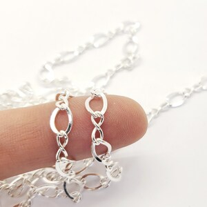 May include: A close-up of a silver chain with oval and figure-eight links. The chain is held by a finger against a white background. The chain is a bright, shiny silver colour.