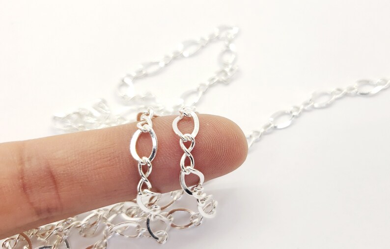 May include: A close-up of a silver chain with oval and figure-eight links. The chain is held by a finger against a white background. The chain is a bright, shiny silver colour.