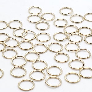 May include: A large quantity of gold jump rings, a type of jewelry finding, are scattered on a white surface.