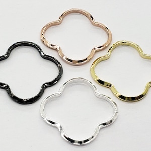 May include: Four openwork clover-shaped charms in various metallic finishes. The charms include black, rose gold, silver, and gold. The charms are likely for jewelry making or other craft projects.