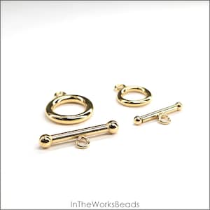 May include: Close-up of gold-colored toggle clasps and rings. The clasps have a bar with rounded ends and a small loop. The rings are circular with a small loop. The clasps and rings are on a white background.