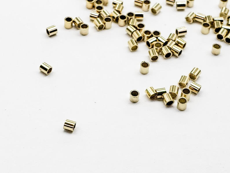 May include: A scattering of gold-colored metal crimp beads on a white surface. The beads are small and cylindrical in shape.