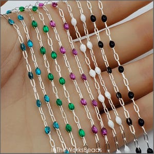 May include: Close-up of multiple strands of silver chain with colorful, oval-shaped beads. The beads are in various colors, including blue, green, purple, white, and black. The chains are held in a hand, with the text "InTheWorksBeads" visible at the bottom.