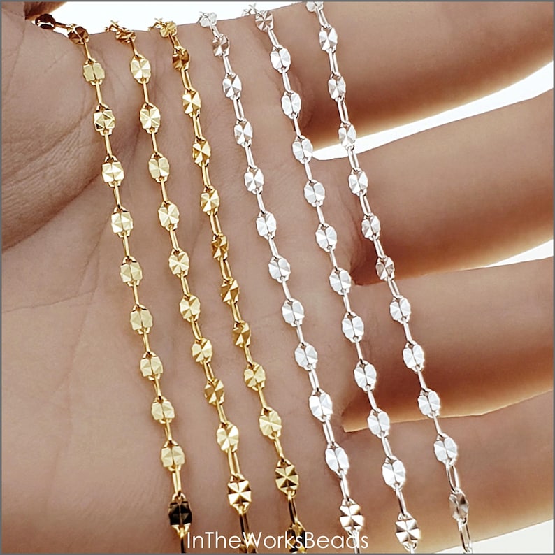 May include: Four chains of different colors, gold, silver, and two shades of gold, are shown on a hand. The chains are made of small, flat, oval-shaped links with a diamond-shaped pattern.
