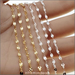 May include: Three strands of chain, one gold, one silver, and one white gold, are shown on a hand. The chains are made of small, oval-shaped links with a faceted finish.