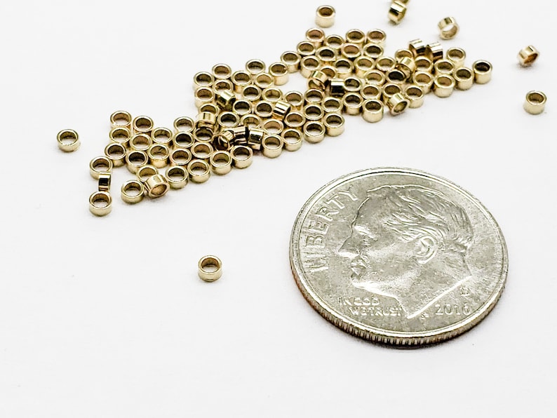 May include: A pile of gold jump rings, some of which are scattered around a US dime coin. The jump rings are small and round, and they are used for jewelry making.