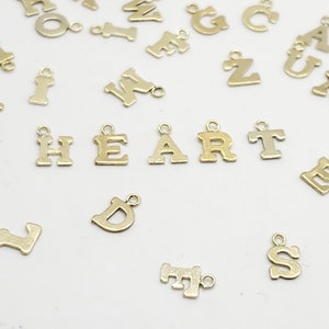 May include: A collection of small, gold-colored metal letter charms scattered on a white surface. The word "HEART" is spelled out with individual charms. Other letters are also visible, suggesting a variety of personalization options for jewelry or crafts.