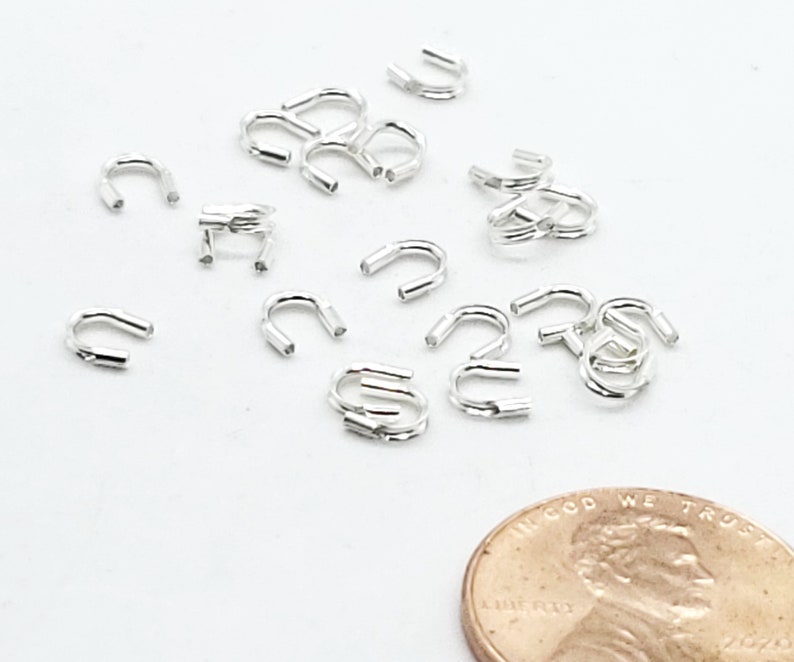 Op de afbeelding: Een close-up van een stapel zilverkleurige metalen krimpkralen. De kralen zijn klein en hebben een U-vorm. Een cent is opgenomen voor schaal.