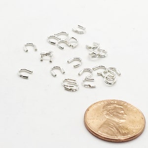 Op de afbeelding: Een close-up van een groep zilverkleurige metalen springringen, verspreid over een wit oppervlak. Een Amerikaanse penny is opgenomen voor schaal.