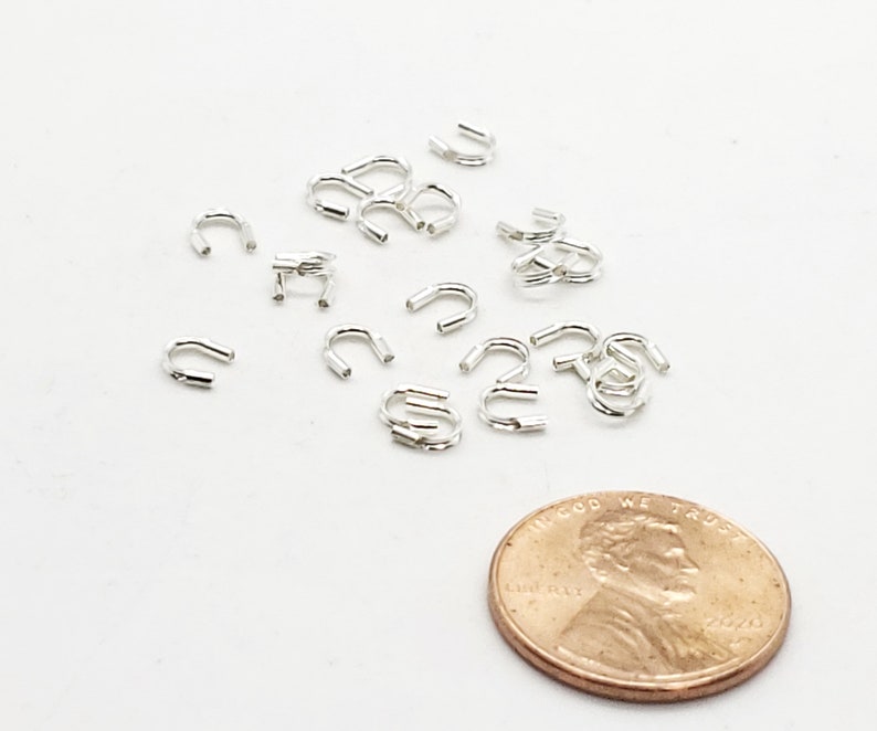 Op de afbeelding: Een close-up van een groep zilverkleurige metalen springringen, verspreid over een wit oppervlak. Een Amerikaanse penny is opgenomen voor schaal.