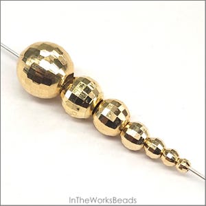May include: A set of six gold-toned round beads with a faceted, textured surface. The beads are arranged in descending size order, with the largest bead at the top and the smallest bead at the bottom.