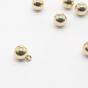 May include: A group of six gold-toned round buttons with a small loop on the top. The buttons are scattered on a white surface.