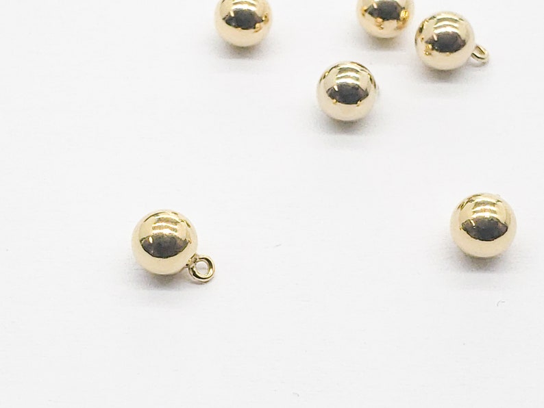 May include: A group of six gold-toned round buttons with a small loop on the top. The buttons are scattered on a white surface.