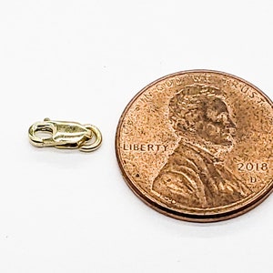Puede incluir: Un cierre de metal dorado para un collar o pulsera, mostrado junto a un centavo de d&oacute;lar estadounidense para comparar el tama&ntilde;o.