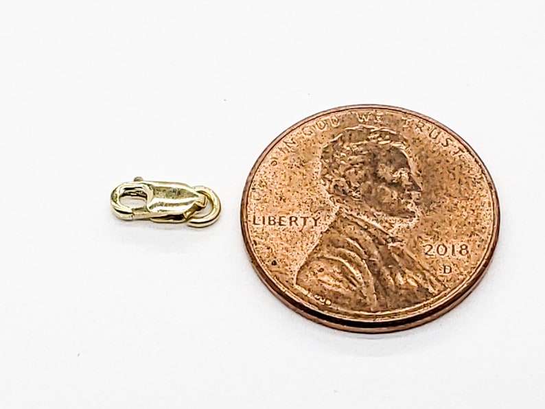 Puede incluir: Un cierre de metal dorado para un collar o pulsera, mostrado junto a un centavo de d&oacute;lar estadounidense para comparar el tama&ntilde;o.