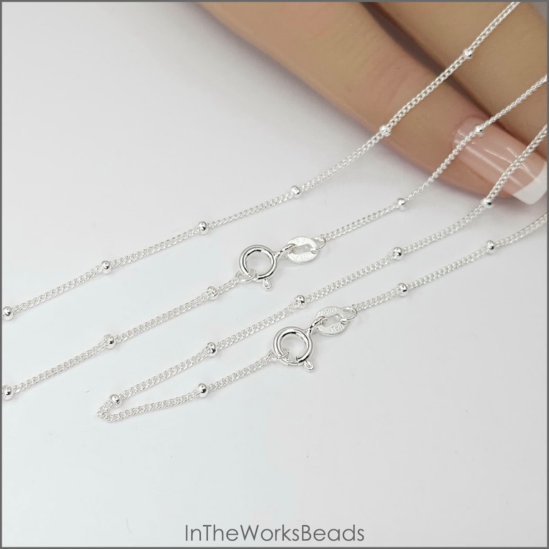 May include: Two silver chains with small round beads. The chains are on a white surface and are laid out in a parallel fashion. The chains are both closed with a clasp.