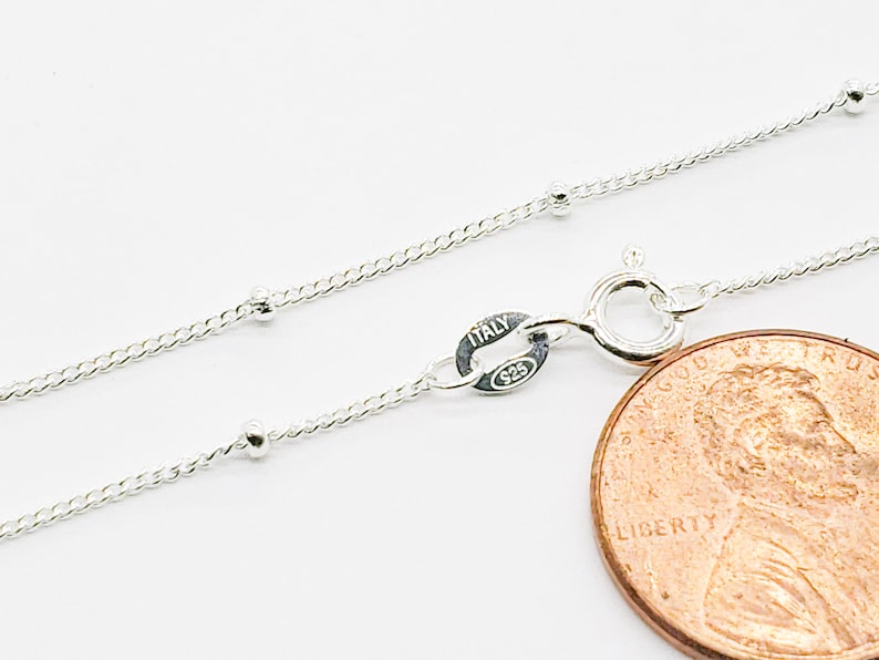 May include: A silver chain necklace with small round beads. The chain is shown next to a US penny for size comparison. The clasp is marked "Italy 925".