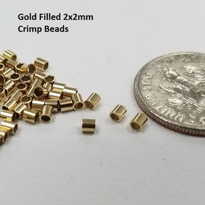 May include: Gold filled 2x2mm crimp beads next to a US dime for size comparison. The beads are small and round, with a hole in the center. They are ideal for jewelry making.