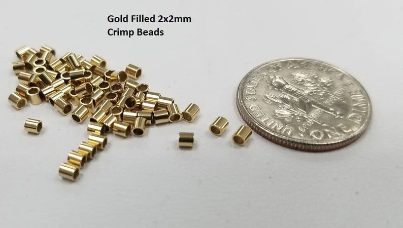 May include: Gold filled 2x2mm crimp beads next to a US dime for size comparison. The beads are small and round, with a hole in the center. They are ideal for jewelry making.