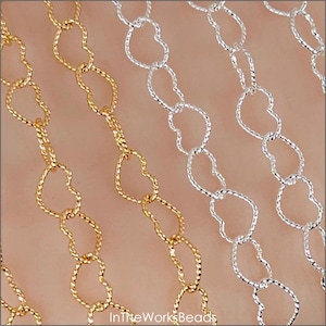 May include: Close-up of gold and silver heart-shaped chain links. The chains are arranged diagonally, with the gold chain on the left and the silver chain on the right. The chains are textured and appear to be made of metal. The text "InTheWorksBeads" is visible at the bottom.