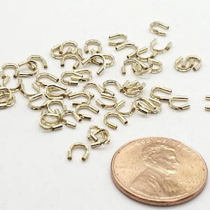 May include: A large quantity of small gold-colored metal jump rings, scattered on a white surface. A US penny is included for scale.