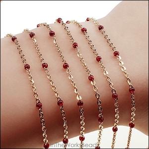 May include: Several gold-coloured chain bracelets, each adorned with small red beads. The bracelets are shown on a person's arm. The image is well-lit, showcasing the jewellery's details. The bracelets appear delicate and elegant.