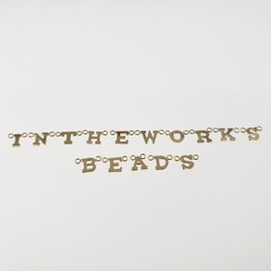 Puede incluir: Cuentas de letras doradas que deletrean "IN THE WORKS" y "BEADS" sobre un fondo blanco.