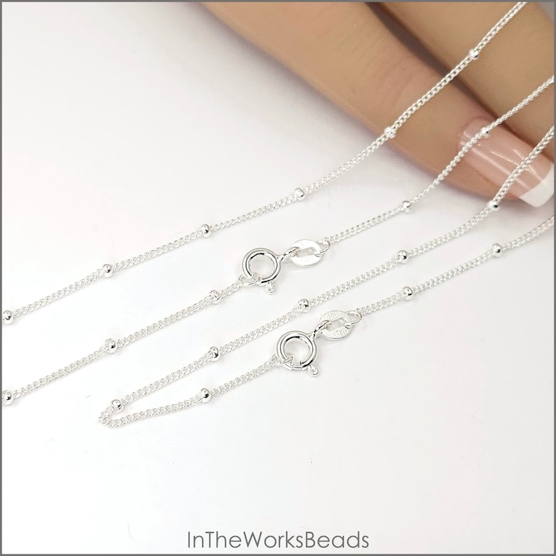 May include: Two silver bead chains with lobster clasps. The chains are layered on top of each other, with the longer chain on top. The chains are on a white background.