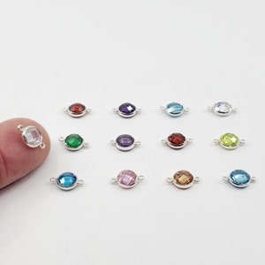 May include: Twelve silver connector charms with round faceted gemstones in various colors, including clear, green, red, purple, blue, and light brown. The charms have two loops on either side for attaching to jewelry.