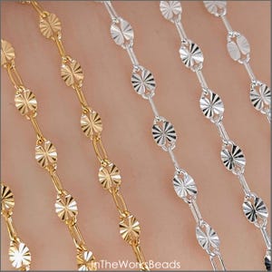 May include: Close-up of gold and silver-colored chain necklaces. The chains feature a repeating pattern of oval-shaped links with a starburst design. The chains are displayed on a neutral background.